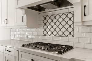 Kitchen with range hood, light stone countertops, stainless steel gas stovetop, backsplash, and white cabinets