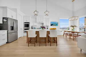 Two tone kitchen featuring suspended lighting, stainless steel appliances, a kitchen island with sink, light wood-style flooring, and a breakfast bar