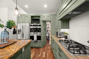 Kitchen featuring green cabinetry, stainless steel appliances, light stone counters, dark wood finished floors, and decorative light fixtures