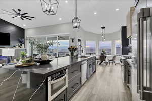 Kitchen with vaulted ceiling, stainless steel appliances, a ceiling fan, light wood finished floors, and an island with sink