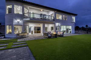 Back of house at night featuring ceiling fan, a balcony, stucco siding, and a patio
