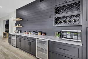 Indoor dry bar with wine cooler, light stone counters, open shelves, light wood finished floors, and stainless steel microwave