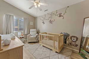 Bedroom with a nursery area, light wood finished floors, ceiling fan, and vaulted ceiling