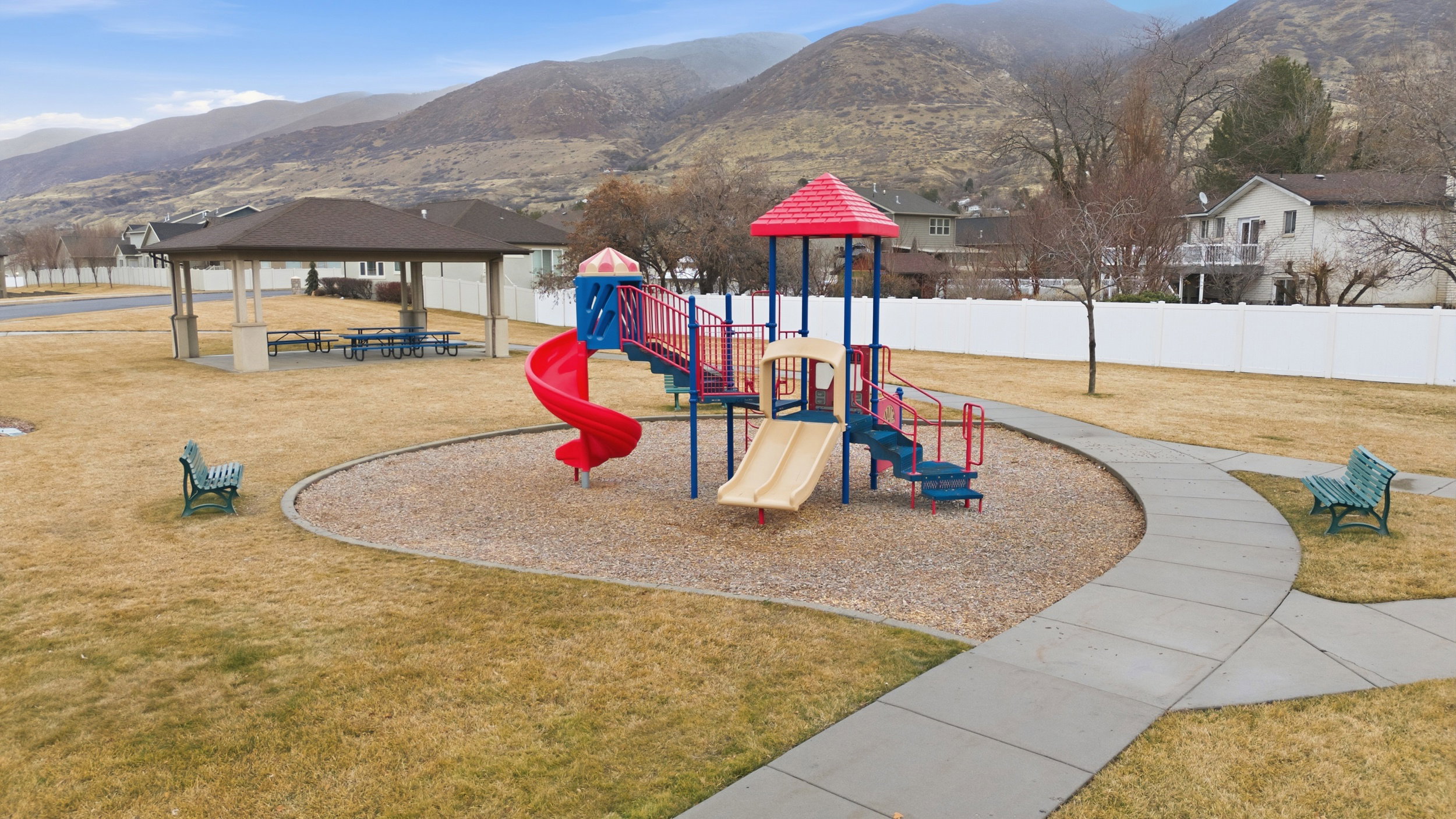 Community playground featuring a multi-level play structure with slides and steps, a covered picnic area with multiple tables, and paved walking paths