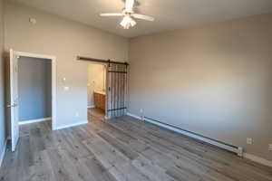 Unfurnished bedroom featuring baseboard heating, connected bathroom, a barn door, light wood finished floors, and a ceiling fan