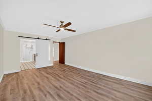 Unfurnished bedroom featuring a barn door, crown molding, wood finished floors, a ceiling fan, and connected bathroom