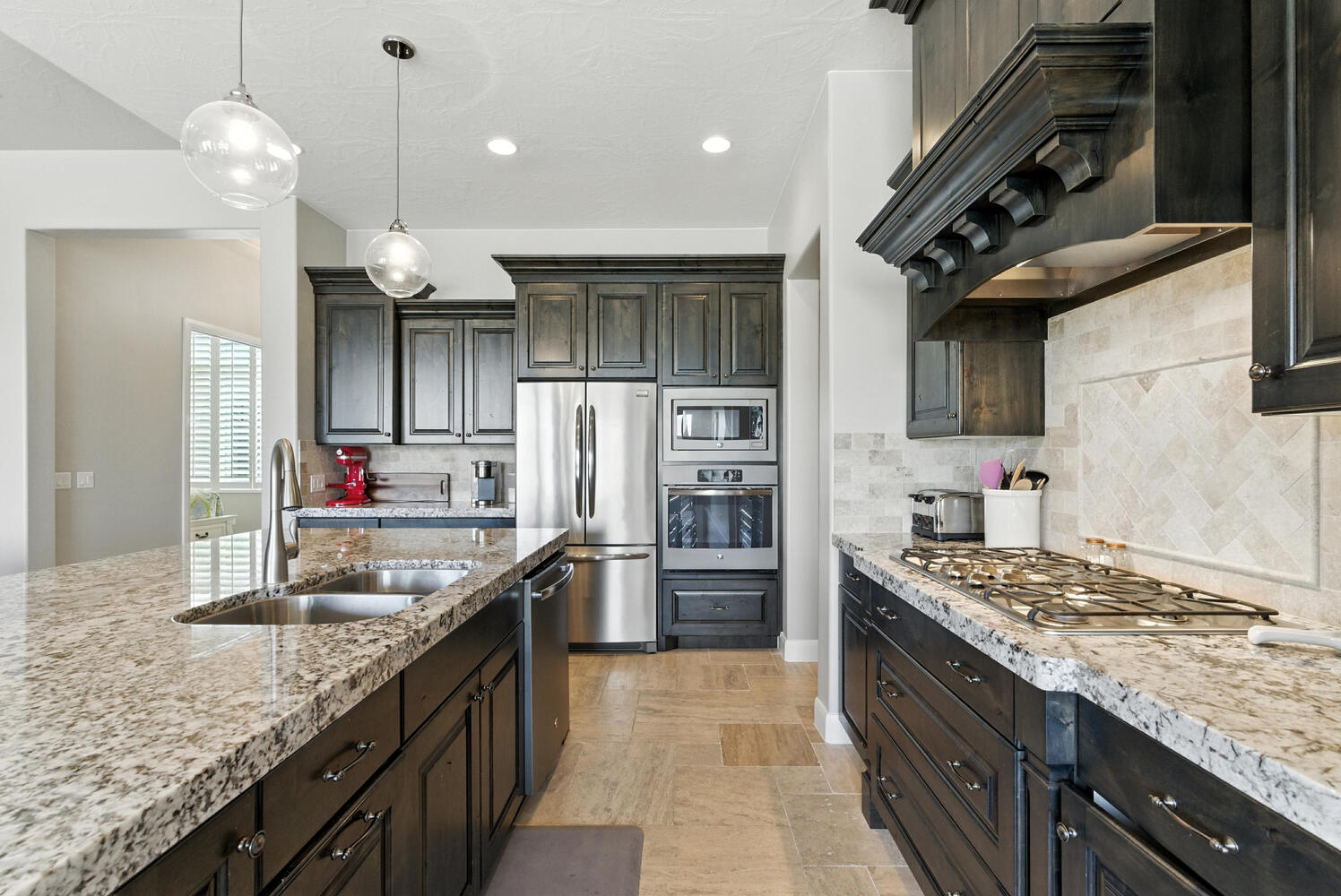 Kitchen featuring stainless steel appliances, pendant lighting, backsplash, light stone counters, and dark wood finish cabinetry