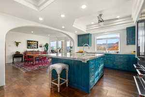 Kitchen featuring blue cabinets, light stone counters, stainless steel fridge, an island with sink, and arched walkways