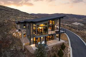 Back of house at dusk with a balcony, a mountain view, stone siding, and a patio area