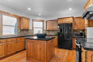 Kitchen featuring black appliances, light wood-style flooring, a kitchen island, ventilation hood, and wood finish cabinets