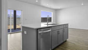 Kitchen featuring gray cabinetry, stainless steel dishwasher, an island with sink, light wood finished floors, and light stone counters