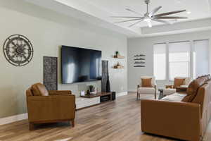Living area with a raised ceiling, light wood finished floors, and a ceiling fan