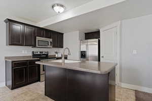 Kitchen with stainless steel appliances, dark wood finish cabinetry, a kitchen island with sink, and light stone countertops