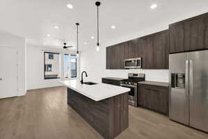 Kitchen featuring stainless steel appliances, dark wood finish cabinets, light wood-type flooring, a kitchen island with sink, and hanging light fixtures