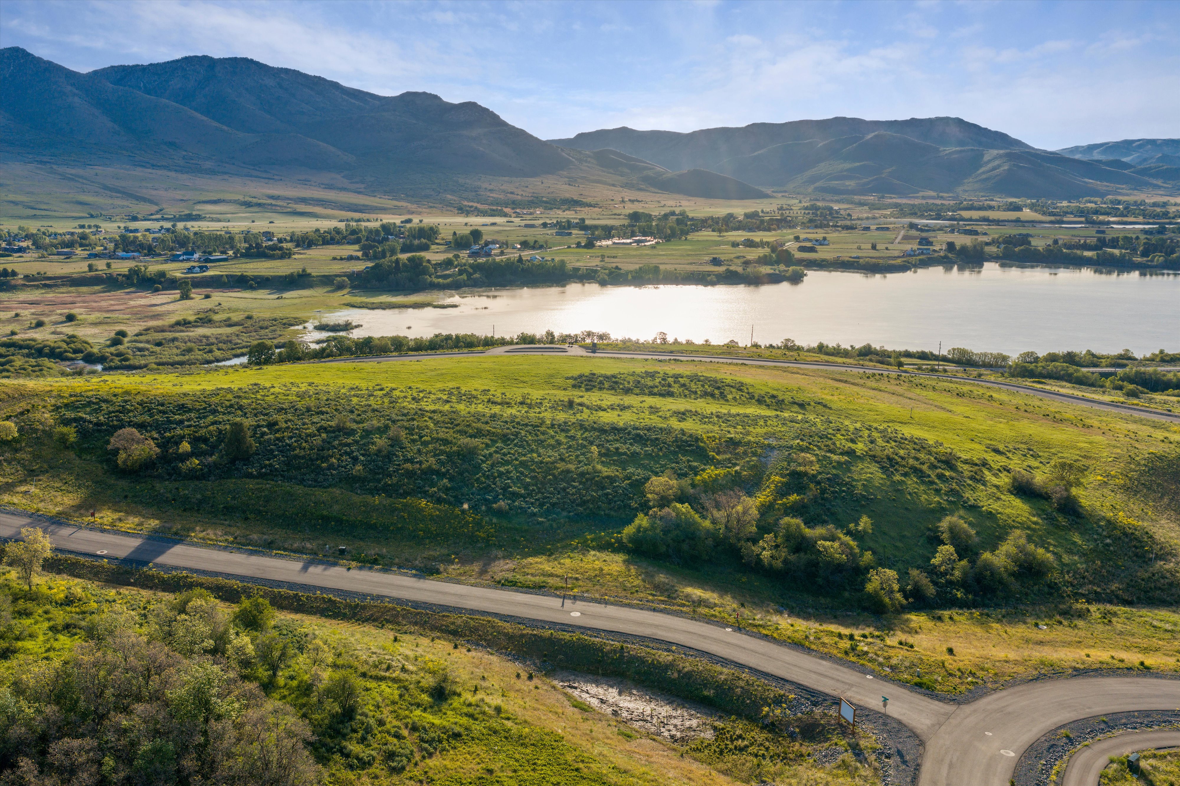 Expansive land parcel featuring a paved roadway, verdant hillsides, and distant water frontage with mountain backdrop