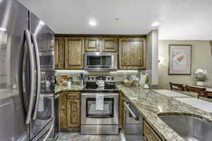 Kitchen with appliances with stainless steel finishes, light stone countertops, brown cabinetry, a textured ceiling, and recessed lighting