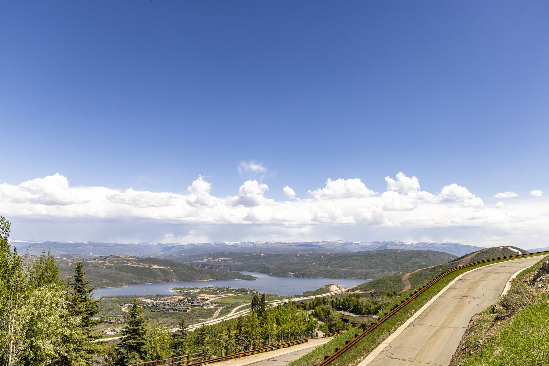 Expansive mountain landscape with a visible lake, distant snow-capped peaks, and a paved road