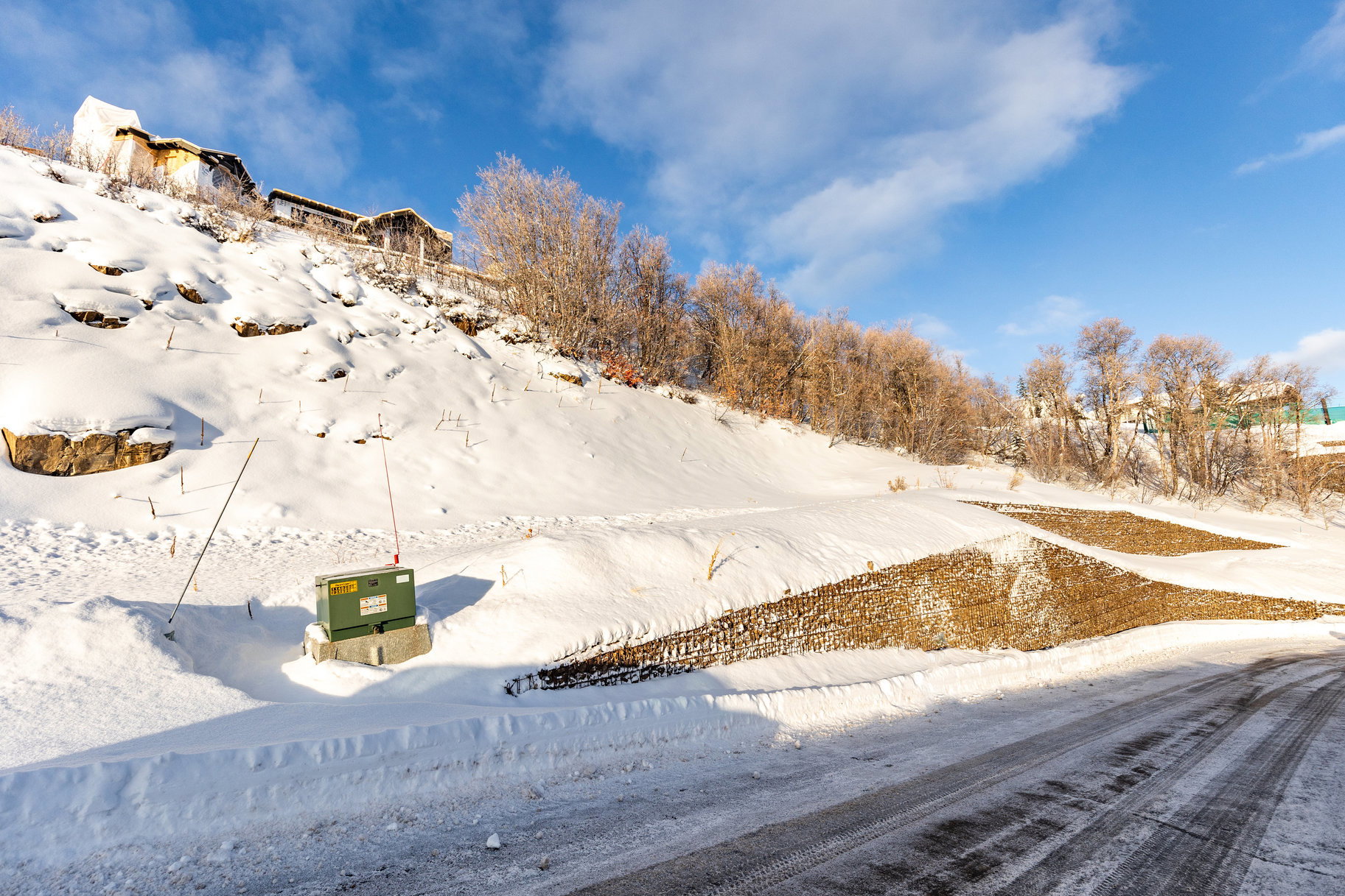 Snow-covered hillside property with visible erosion control measures and a paved road frontage