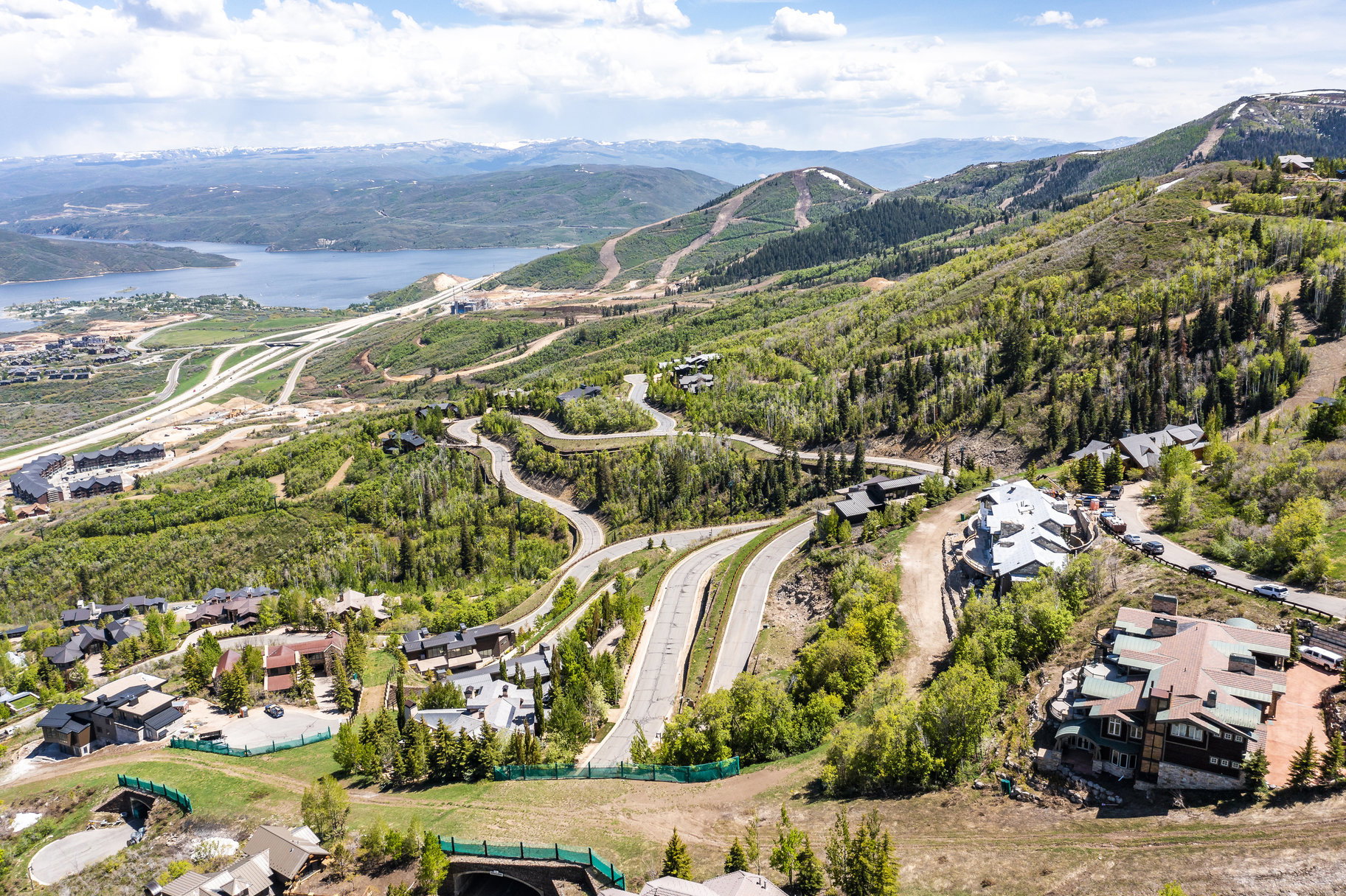 Sweeping mountain landscape featuring established residential properties, winding roadways, abundant mature trees, a distant lake, and snow-capped peaks