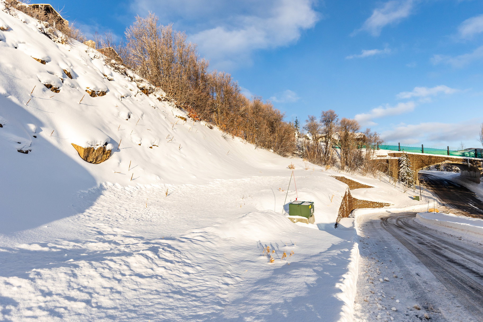 Snow-covered terrain with a winding road alongside, featuring a stone-faced retaining wall and an overhead bridge structure