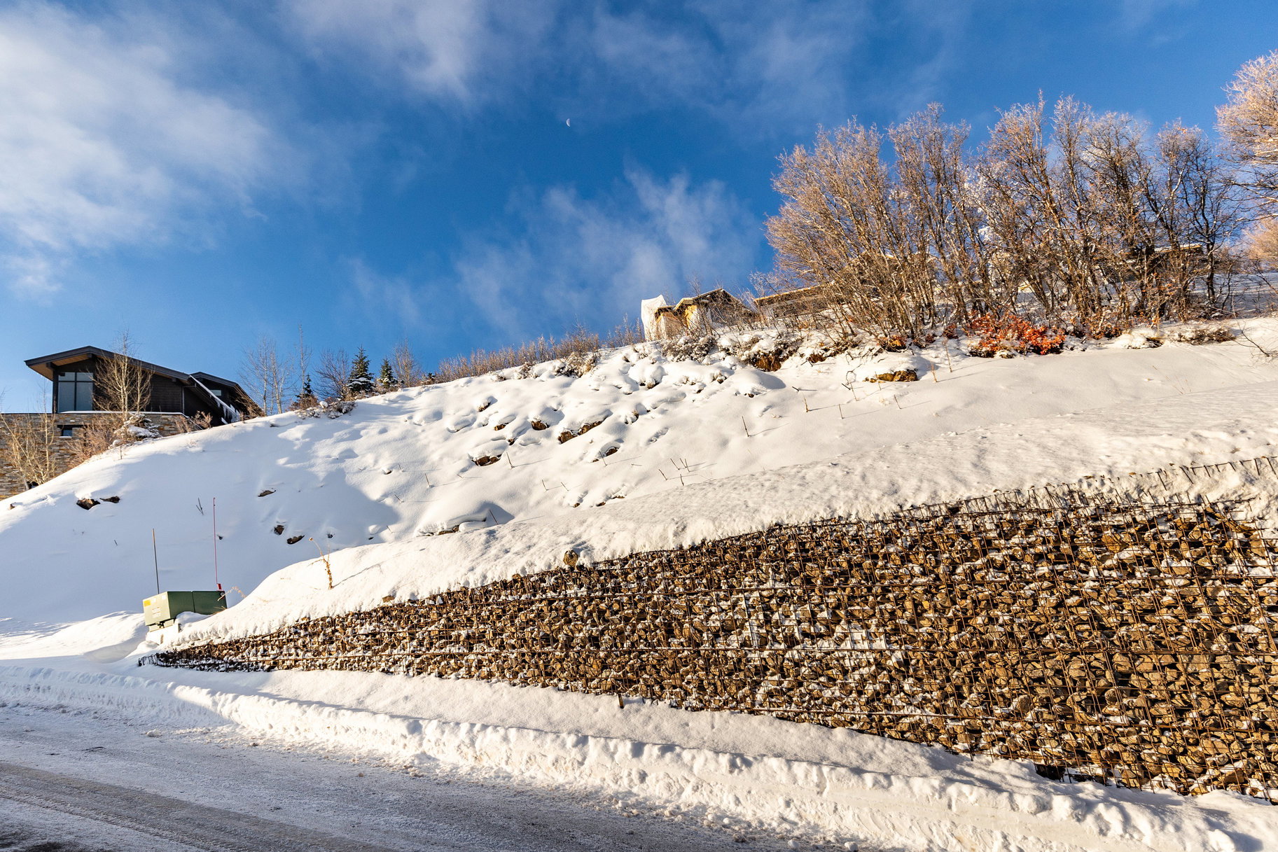 Hillside property featuring a modern architectural design with large windows, a gabion wall retaining structure, and a snow-covered landscape