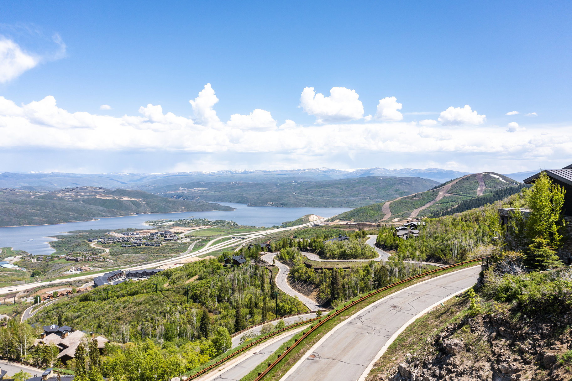 Expansive mountain and lake landscape featuring a winding paved road, distant snow-capped peaks, and scattered residential properties