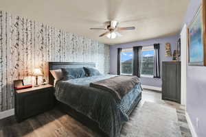 Bedroom with wallpapered walls, wood finished floors, an accent wall, and a ceiling fan
