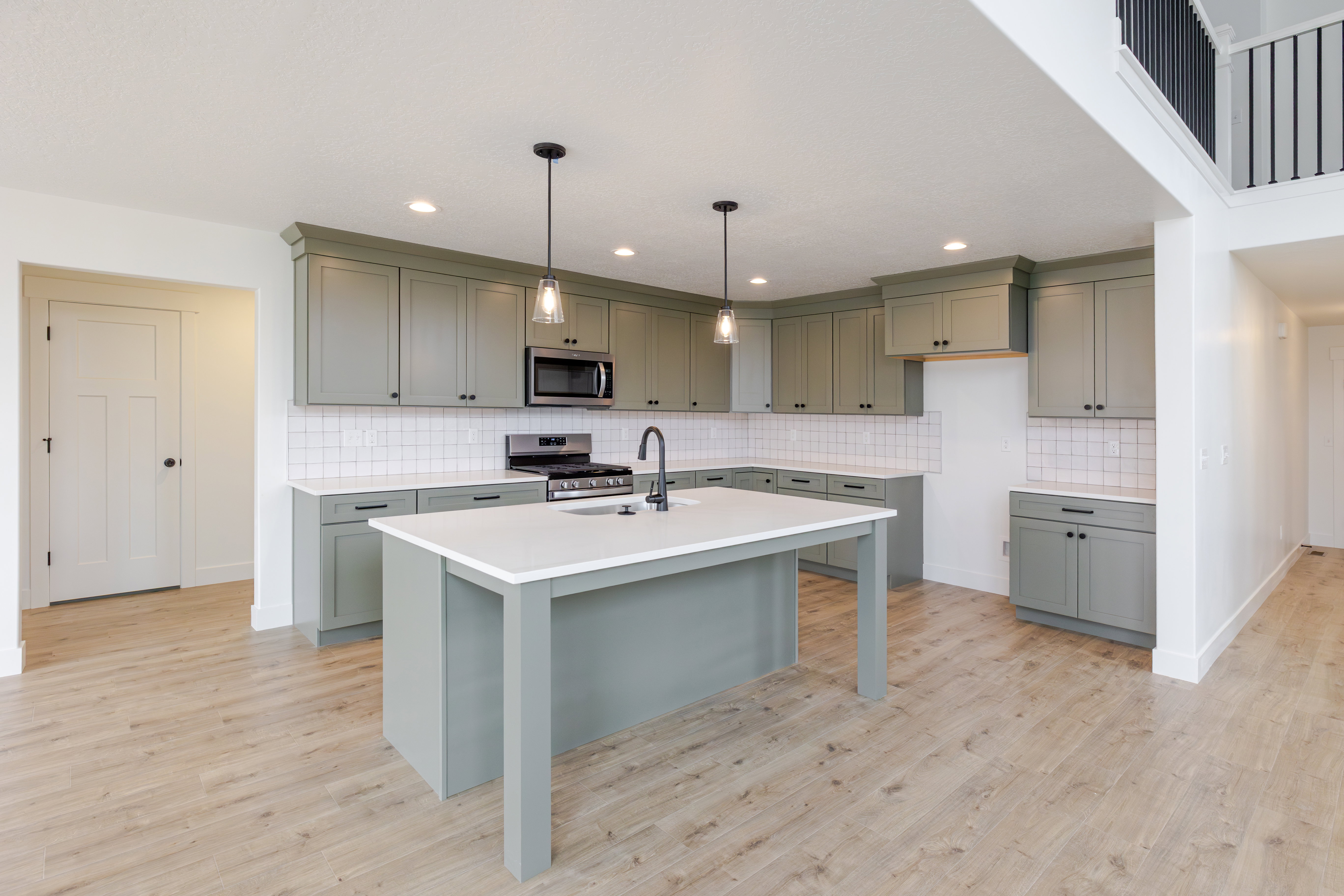 Open-concept kitchen featuring an oversized island with a white countertop, extensive sage green cabinetry, a white subway tile backsplash, stainless steel appliances, and wood-finish flooring throughout