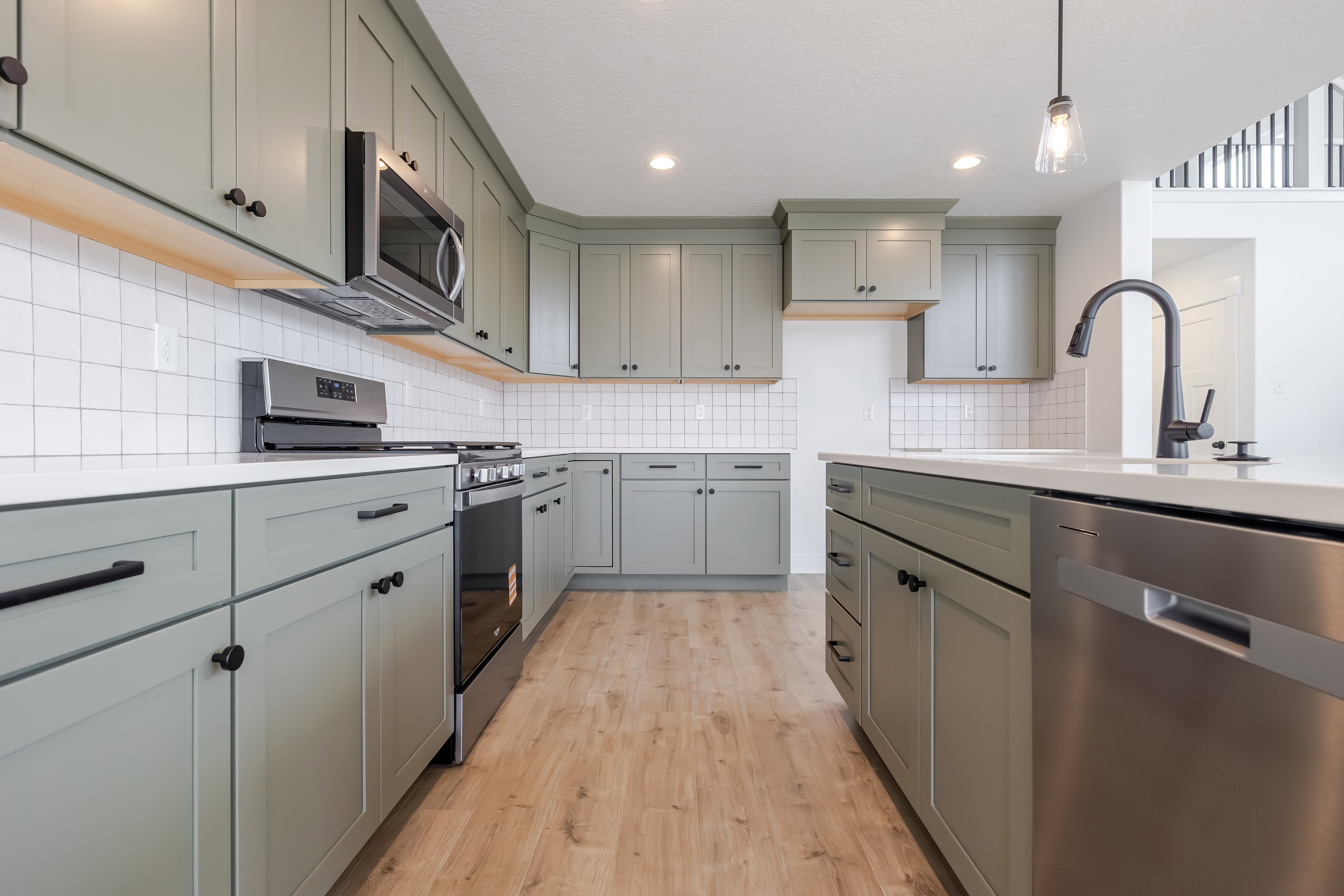 Kitchen featuring light green cabinetry with black hardware, white subway tile backsplash, light-colored countertops, stainless steel appliances, and wood-finish flooring