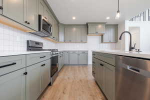 Kitchen featuring light green cabinetry with black hardware, white subway tile backsplash, light-colored countertops, stainless steel appliances, and wood-finish flooring