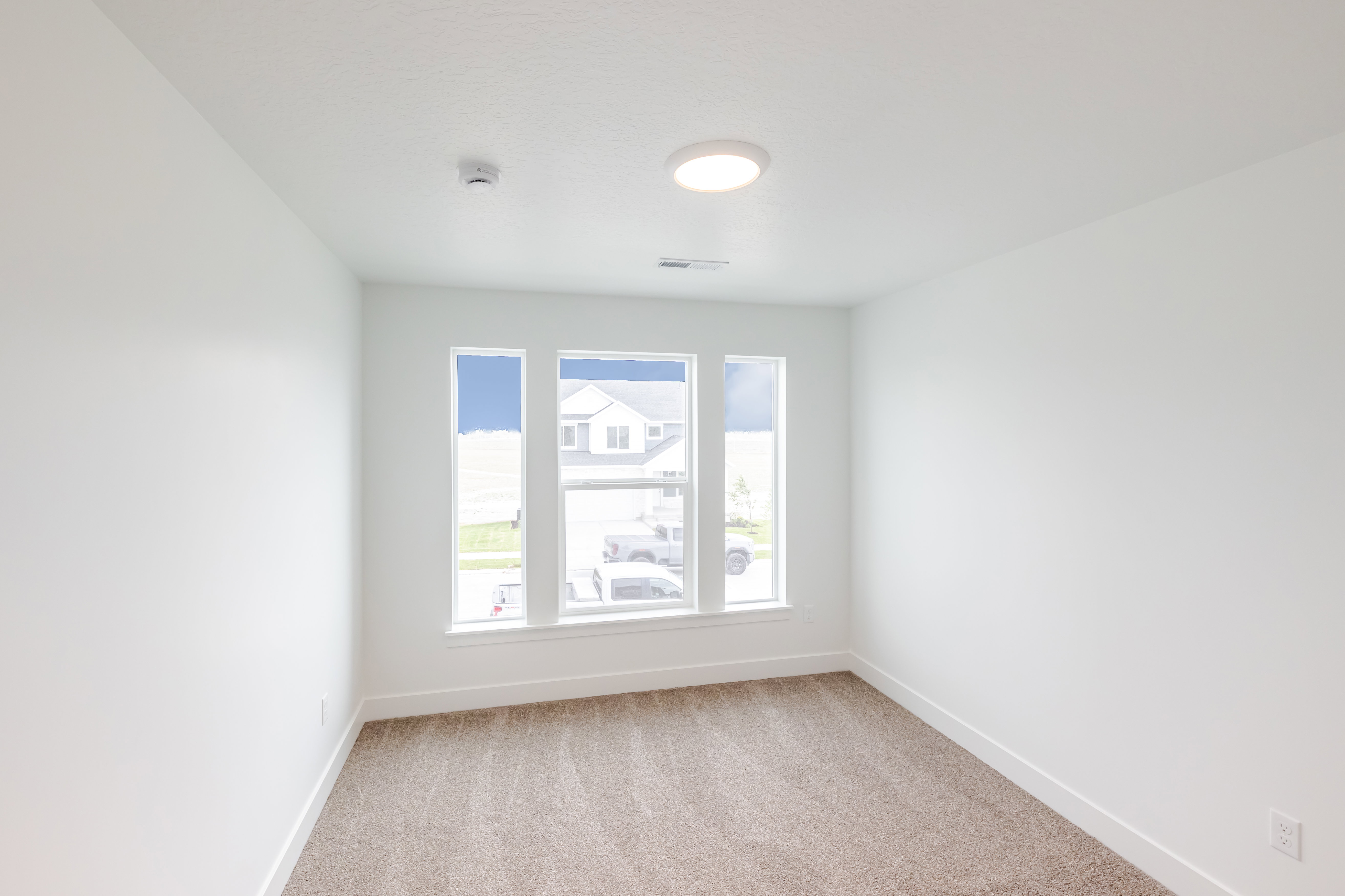 Bright interior space featuring three large windows, neutral-toned carpeting, white walls, and a ceiling-mounted light fixture