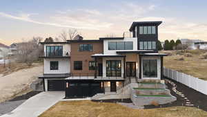 Back of property featuring a balcony, driveway, a standing seam roof, an attached garage, and a patio
