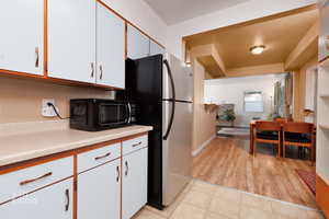Kitchen view into dining and living room area. Dual tone kitchen featuring light countertops, black microwave, light tile patterned flooring, two tone cabinets, and freestanding refrigerator