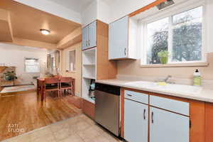 Kitchen view into dining and living room area. Two tone kitchen with light countertops, stainless steel dishwasher, open shelves, two tone cabinetry, and light tile patterned floors