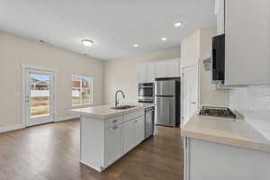 Kitchen featuring a kitchen island with sink, stainless steel appliances, white cabinetry, dark wood-style flooring, and tasteful backsplash