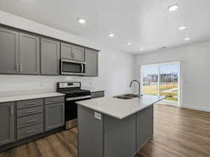 Kitchen with gray cabinetry, stainless steel appliances, dark wood finished floors, a textured ceiling, and a kitchen island with sink