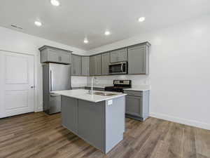 Kitchen featuring gray cabinetry, stainless steel appliances, dark wood finished floors, a kitchen island with sink, and light stone counters
