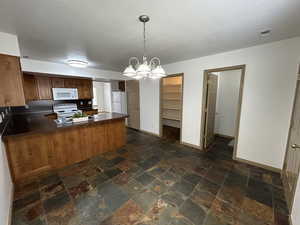Kitchen featuring a peninsula, a chandelier, white appliances, dark countertops, and wood finish cabinets