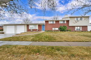 Split level home featuring driveway, brick siding, an attached garage, a chimney, and a front lawn