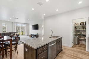 Kitchen with dark wood finish cabinetry, light stone countertops, light wood finished floors, a center island with sink, and stainless steel dishwasher