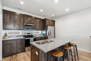 Kitchen featuring stainless steel appliances, light stone counters, dark wood finish cabinetry, a center island with sink, and light wood finished floors