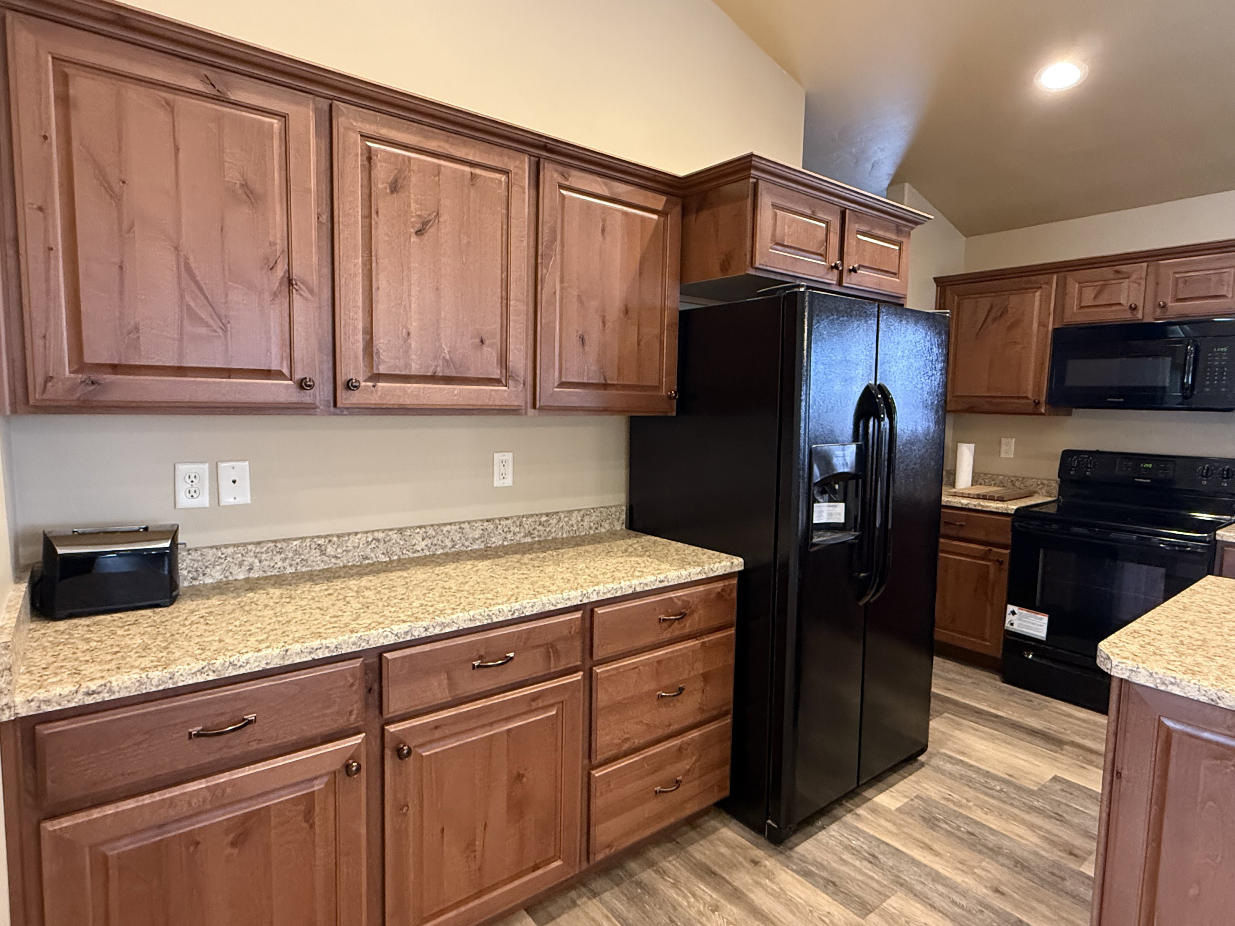 Kitchen featuring black appliances, light wood-style flooring, vaulted ceiling, light stone counters, and wood finish cabinets