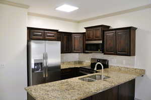Kitchen featuring dark wood finish cabinetry, stainless steel appliances, light stone counters, crown molding, and a peninsula