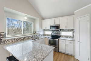 Kitchen with stainless steel appliances, white cabinetry, light stone countertops, backsplash, and lofted ceiling