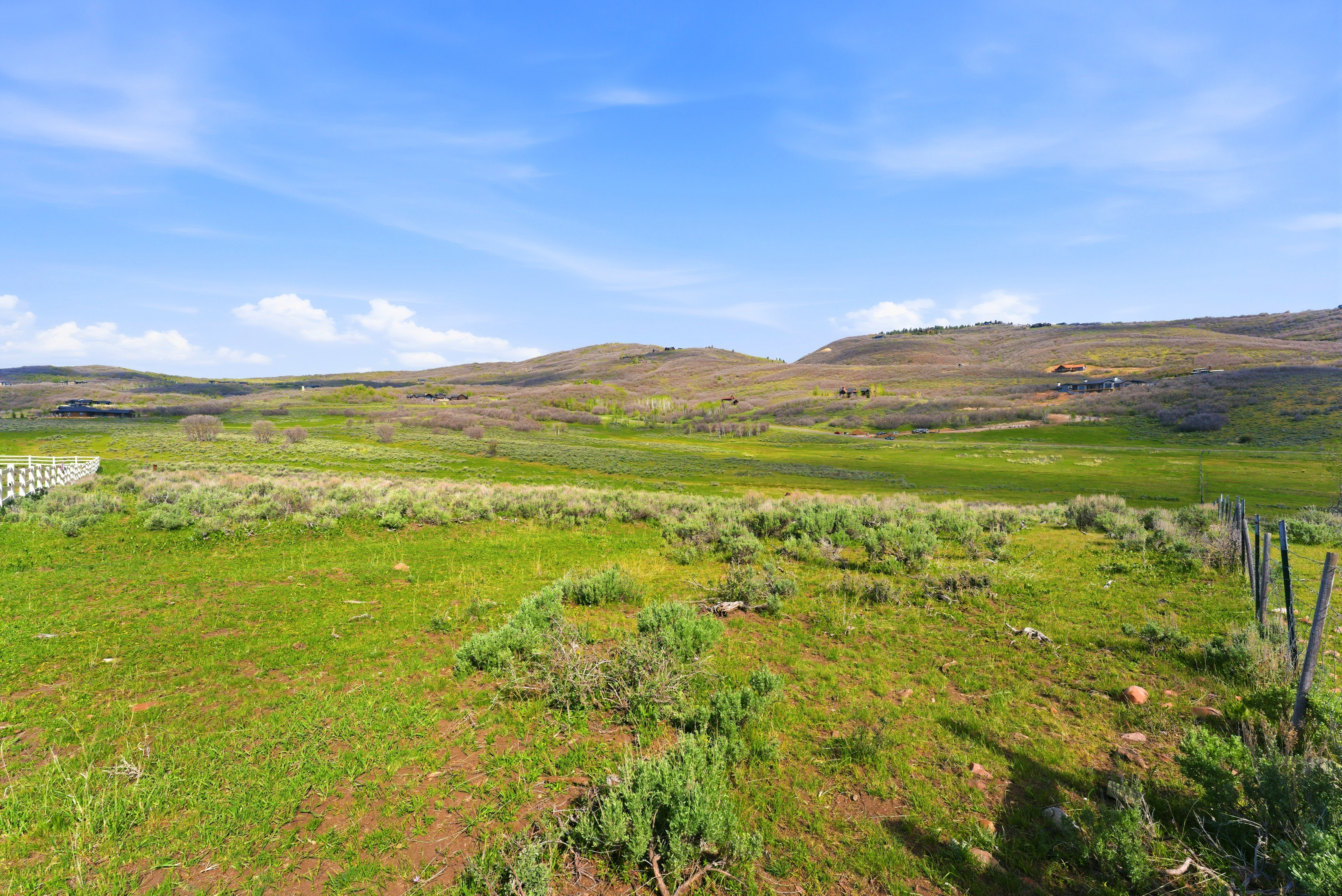 Expansive green landscape featuring rolling hills, scattered low-lying shrubs, and a partial white fence line