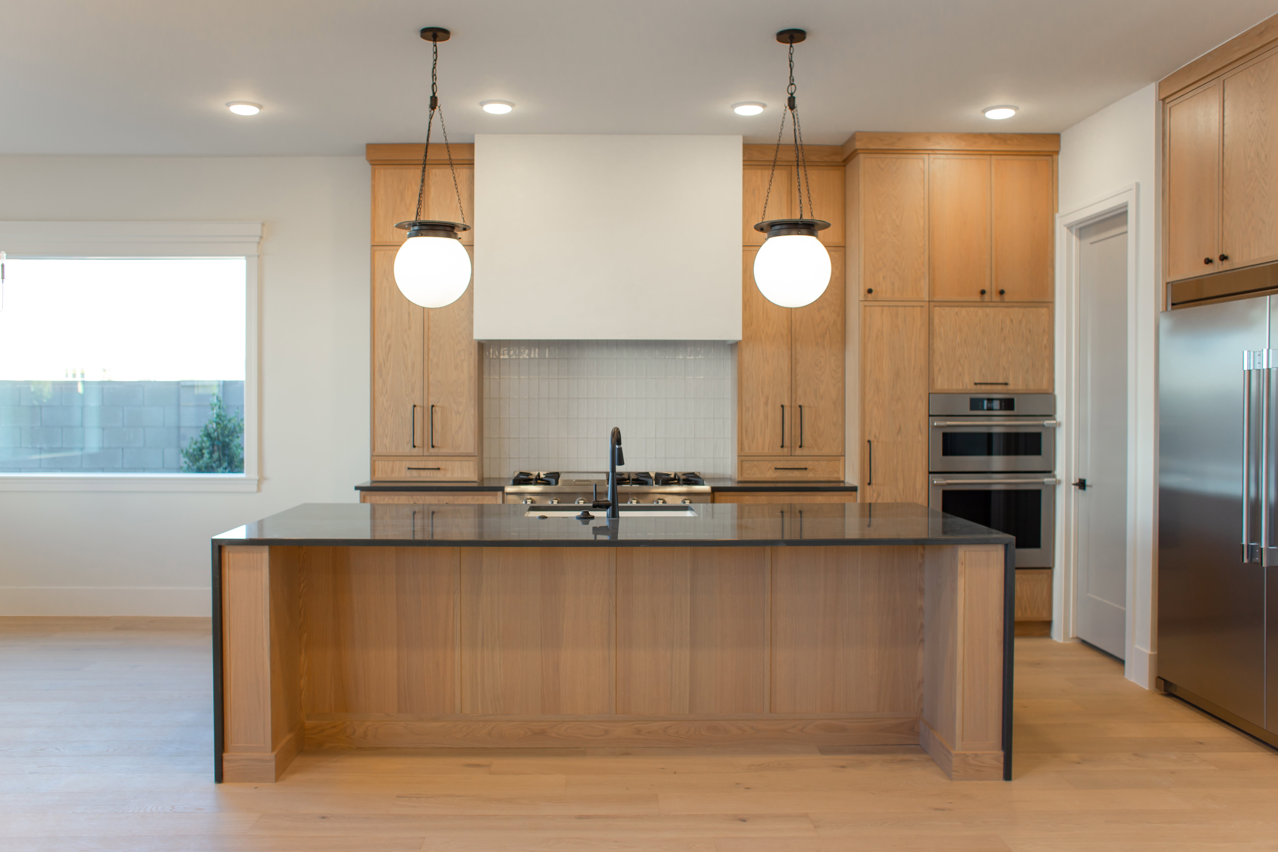 Kitchen featuring stainless steel appliances, light wood-style floors, dark stone counters, decorative light fixtures, and a kitchen island with sink