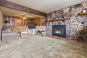 Living room with carpet, a stone fireplace, a textured ceiling, ceiling fan, and wood walls