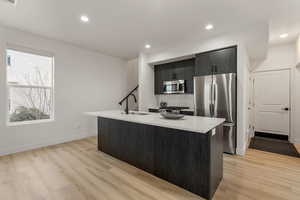 Kitchen featuring stainless steel appliances, an island with sink, light wood-style floors, light stone countertops, and dark cabinetry
