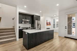Kitchen with a center island with sink, stainless steel appliances, light wood-type flooring, dark cabinets, and tasteful backsplash