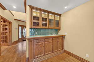 Bar area with brown cabinets, glass insert cabinets, backsplash, light wood finished floors, and tile countertops